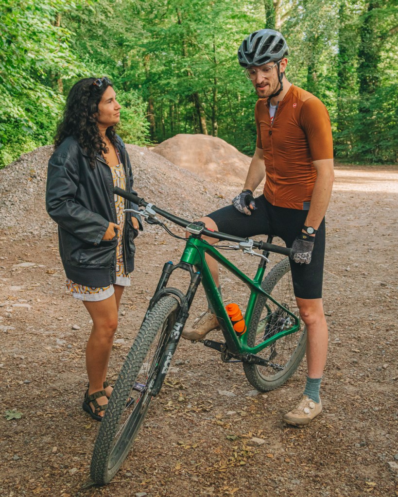 Two people engaging in conversation at a cycling spot in a forest. One person is standing next to a green mountain bike while the other is leaning against a tree.