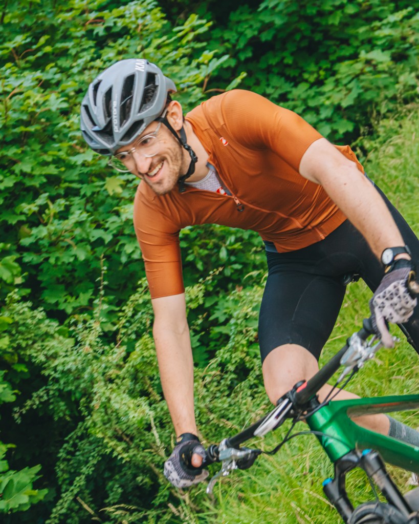 A smiling cyclist wearing a brown jersey and cycling helmet, riding a green mountain bike on a grassy trail surrounded by lush greenery.