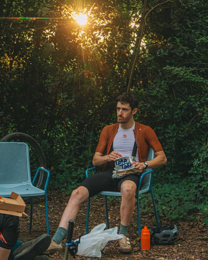 A cyclist sitting on a blue chair in a wooded area, holding a takeaway bag. The sun is setting in the background, creating a warm glow. A pizza box and a water bottle can be seen nearby.