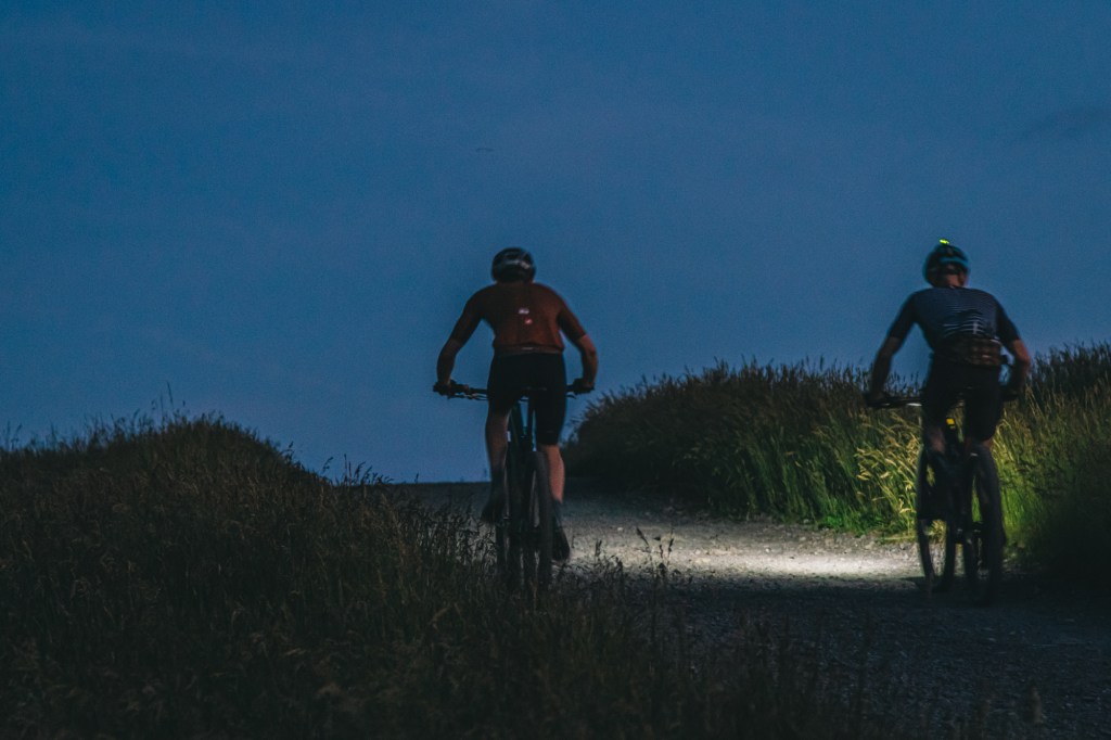 Two cyclists riding on a gravel path at dusk, with their bike lights illuminating the way.