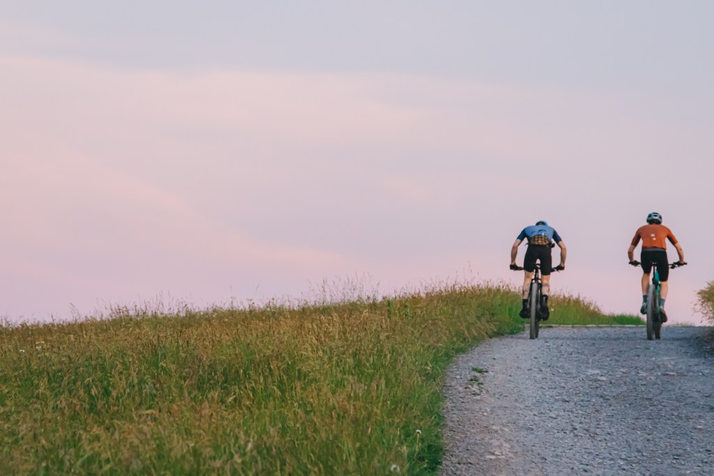 Two cyclists riding up a gravel path surrounded by tall grass, with a pastel sky in the background.