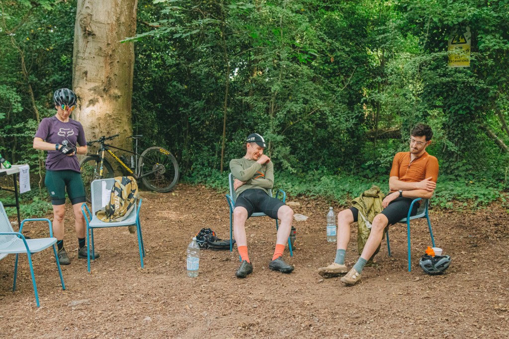 Three cyclists resting in a wooded area, with one person checking equipment, while the others sit on chairs. A bicycle is seen in the background, and water bottles are on the ground.