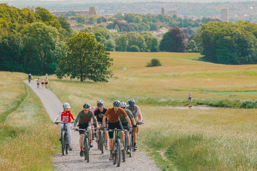 A group of cyclists riding on a gravel path through a lush green landscape, with trees and a distant city skyline in the background.